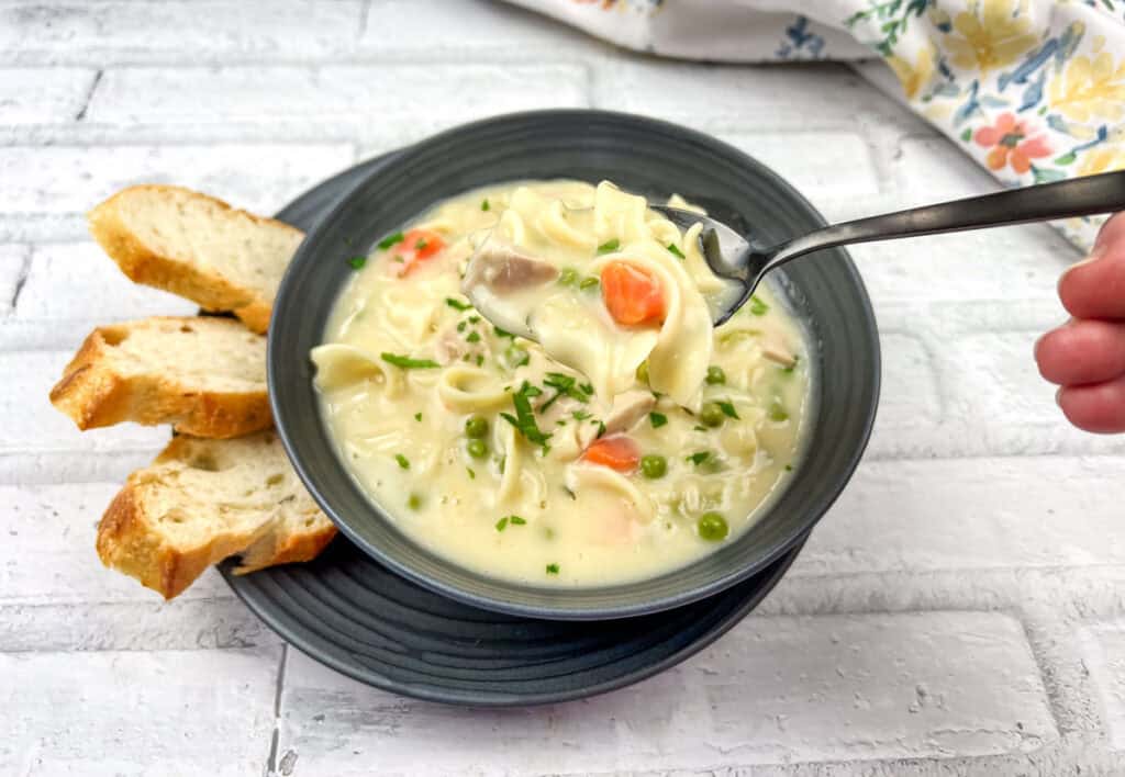 A spoonful of Creamy Chicken Noodle Soup served in a black bowl with slices of bread on a plate.