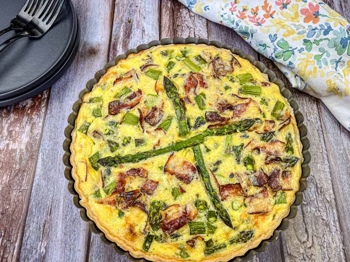 Asparagus & Ricotta Quiche in a round pan on a wooden table, with a floral napkin and black plates with forks nearby.