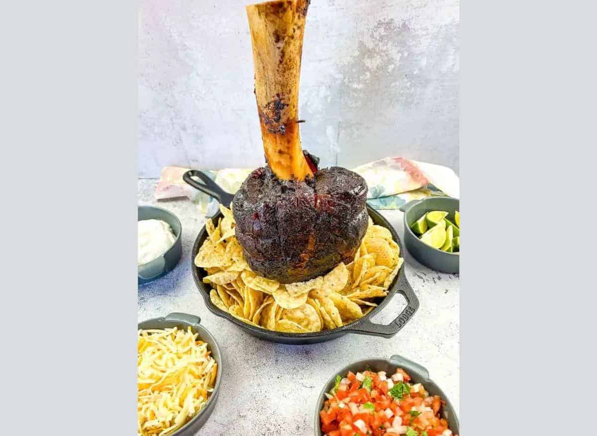 A large roasted meat shank on a bone, displayed upright in a skillet surrounded by nachos, with bowls of lime wedges and sauce nearby.