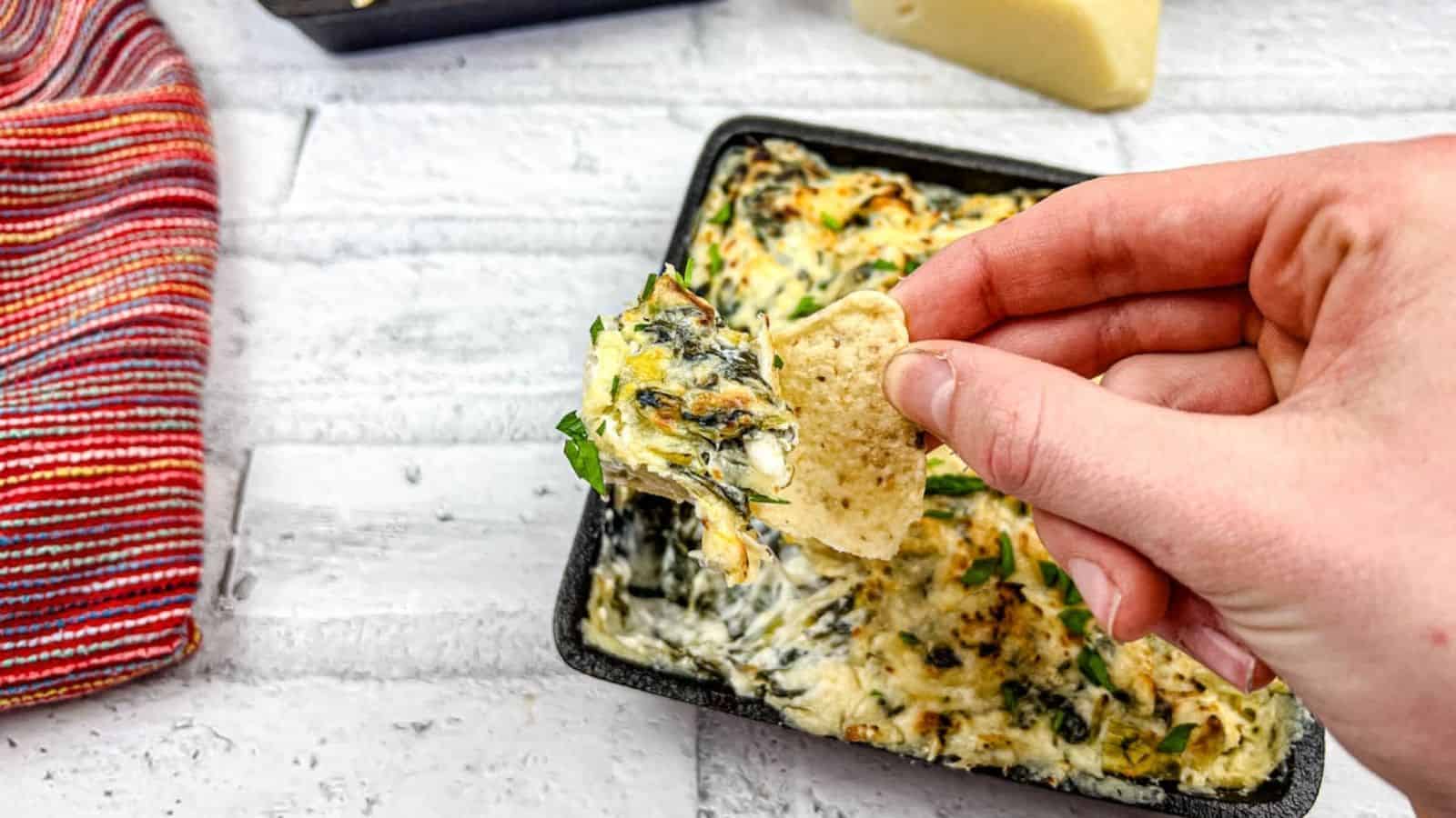 A hand dipping a tortilla chip into a tray of Copycat Applebee’s spinach artichoke dip.