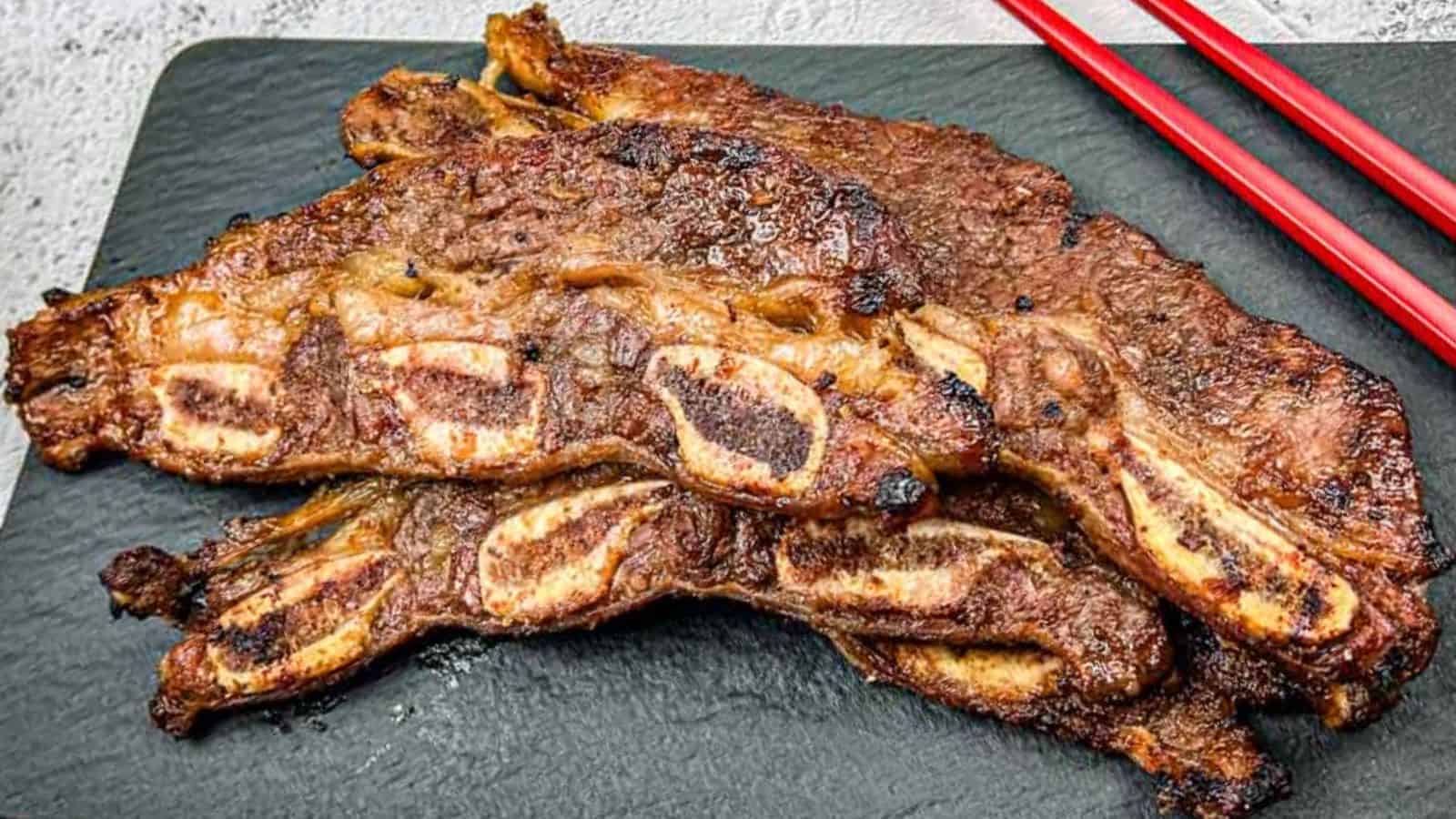 Two Korean galbi ribs on a black slate plate with red chopsticks placed beside them.