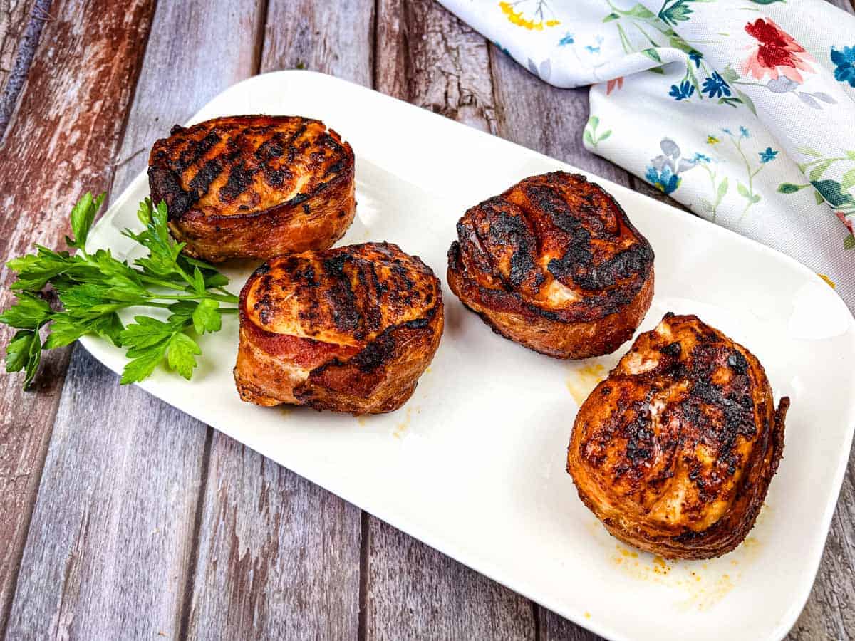 Four bacon-wrapped chicken breasts on a white rectangular plate garnished with sprigs of parsley.