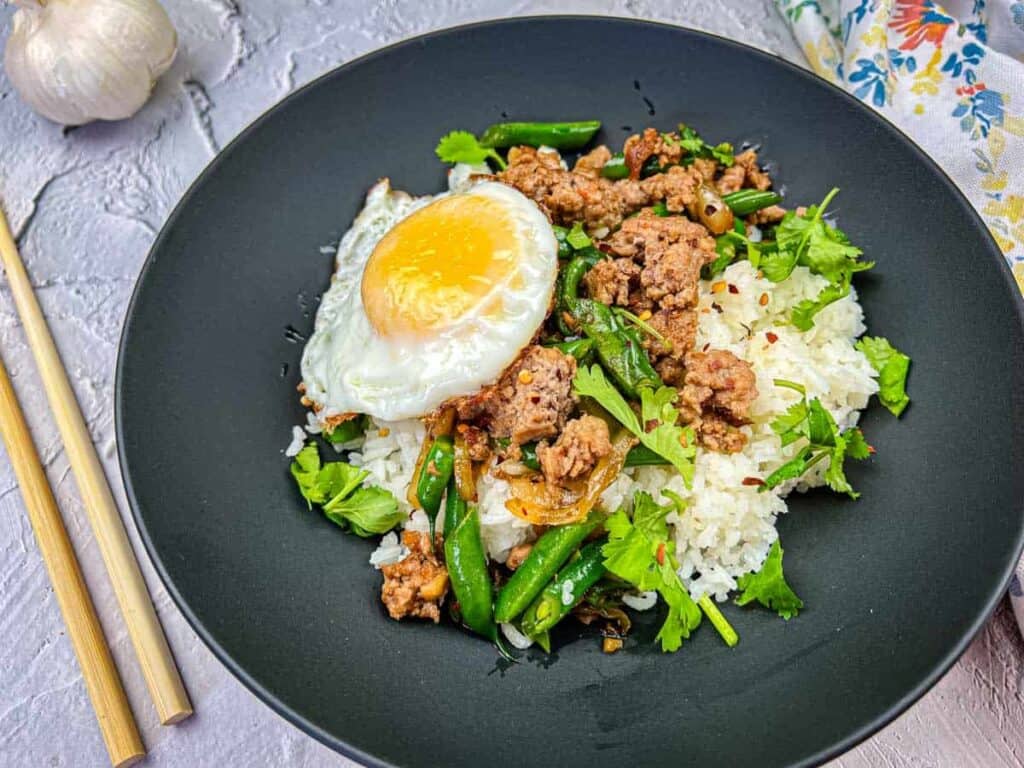 A plate of Thai pork & Green beans with rice, topped with a fried egg, served with chopsticks on the side.