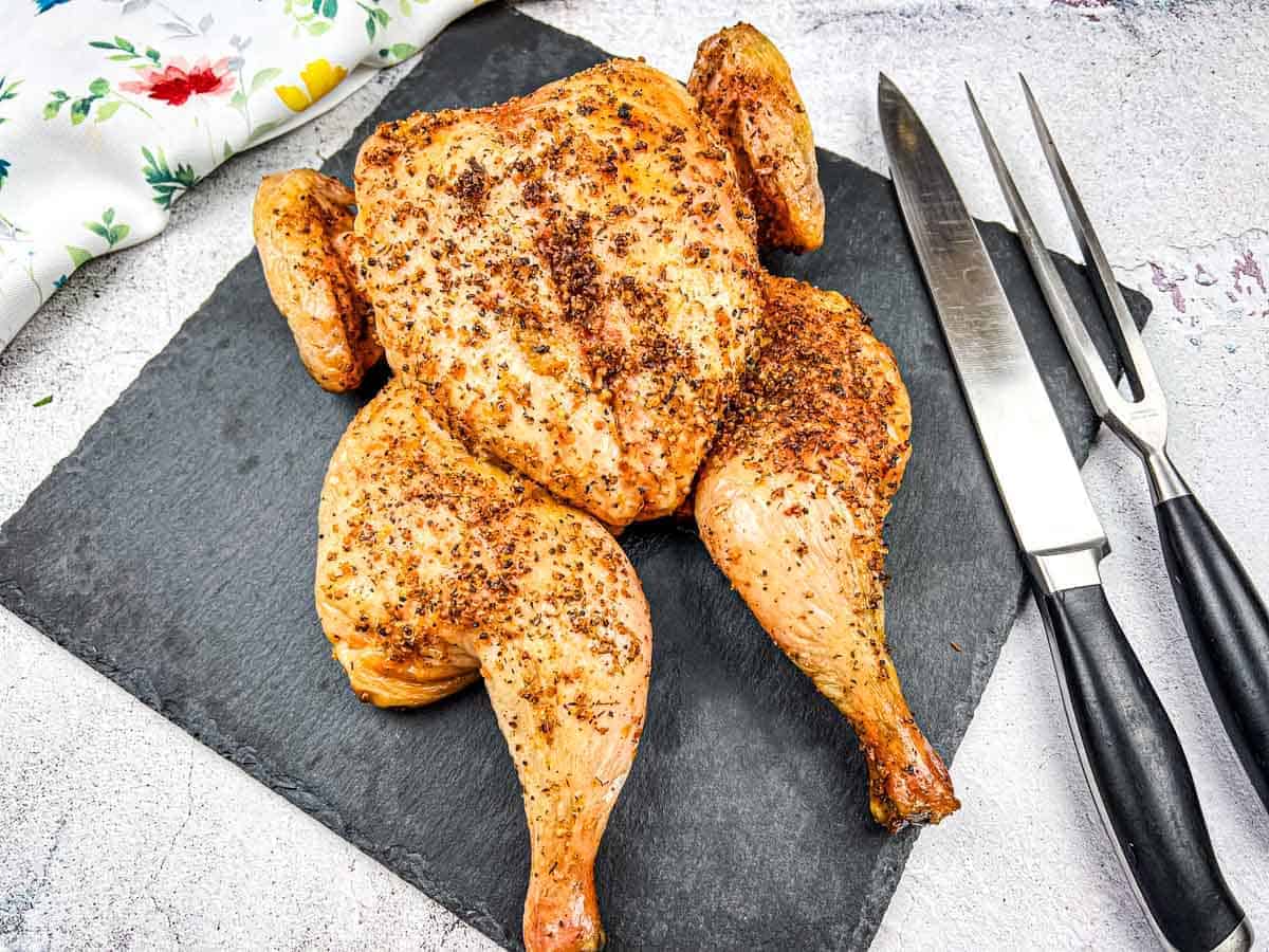 A Traeger smoked spatchcock chicken on a slate board next to a carving knife and fork.