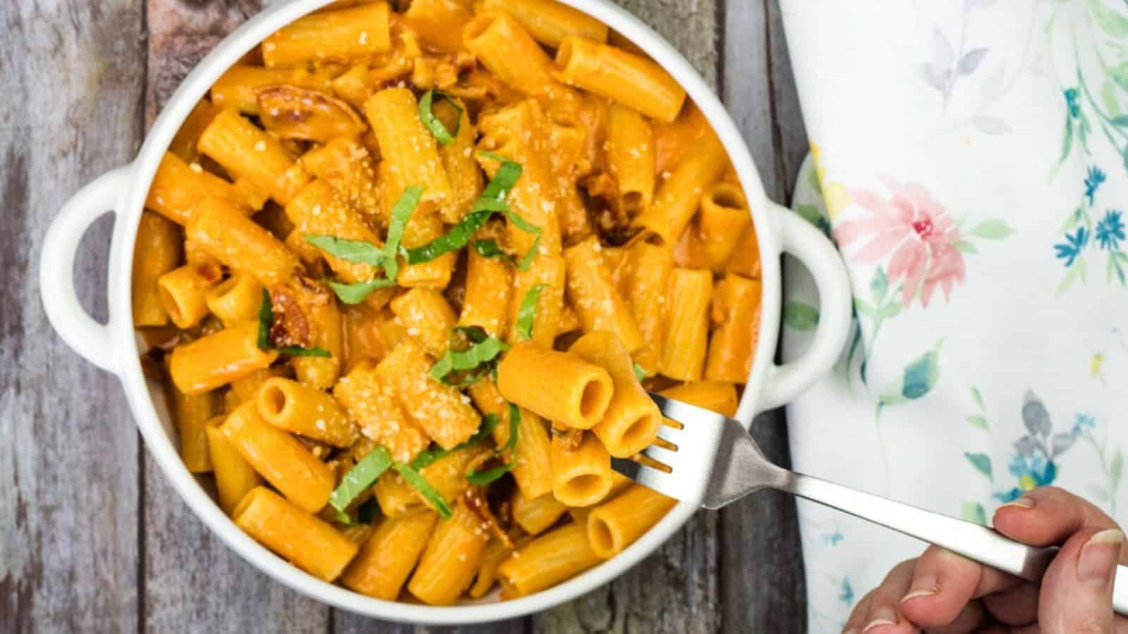 A white bowl of rigatoni pasta with creamy sauce, garnished with cheese and green herbs, is placed on a wooden surface. A hand is holding a fork with a piece of pasta.