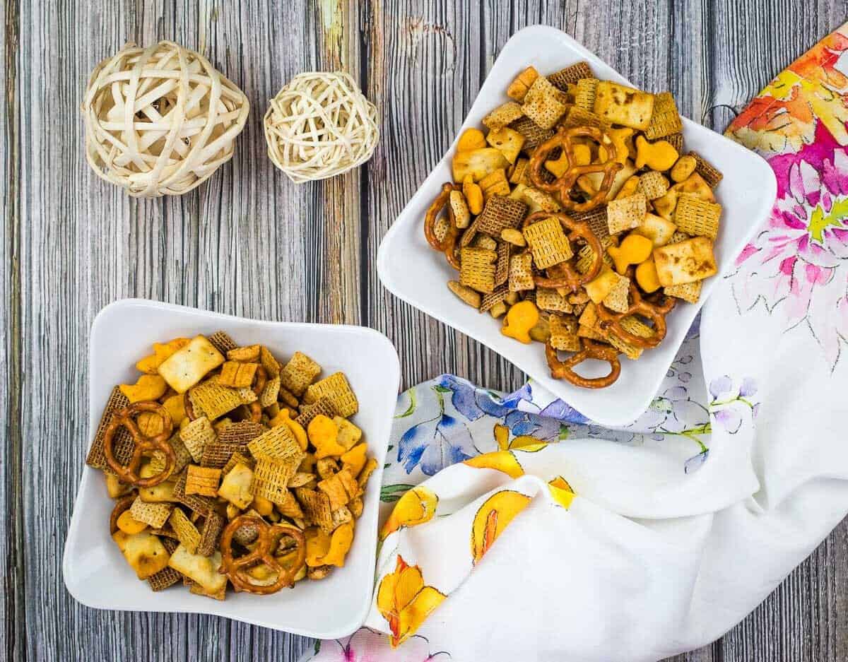 Two bowls of snack mix with pretzels and cereal on a wooden table, accompanied by decorative balls and a colorful napkin.