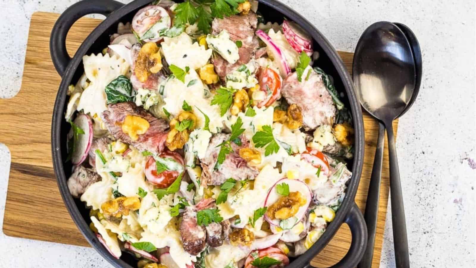 A black bowl filled with a pasta salad containing farfalle, steak slices, radishes, cherry tomatoes, lettuce, nuts, and crumbled blue cheese, garnished with parsley, on a wooden board next to a spoon.