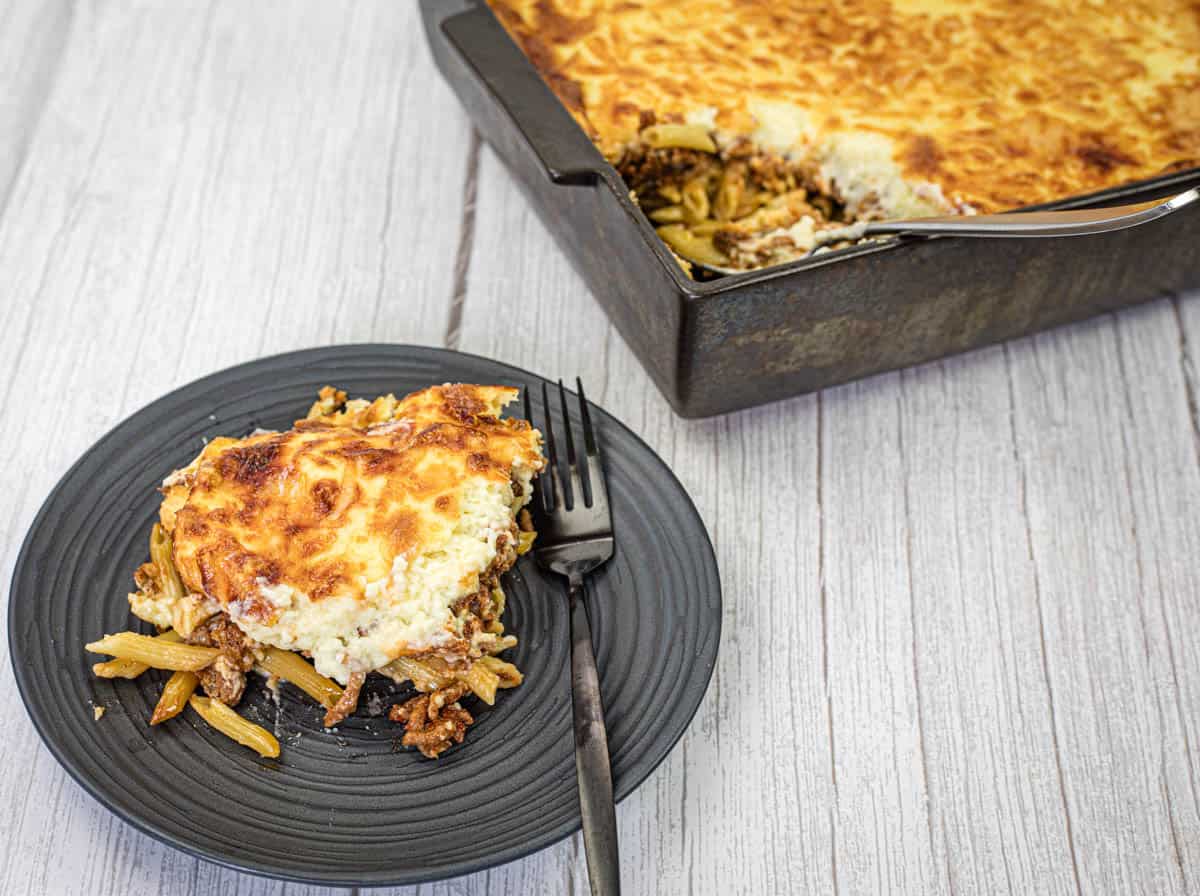 A portion of Pastitsio on a black plate beside the tray of Pastitsio.