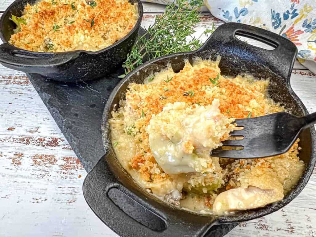 Two cast-iron skillets filled with creamy, baked casserole topped with golden breadcrumbs and garnished with fresh thyme sprigs, set on a wooden surface. A fork rests in one serving.