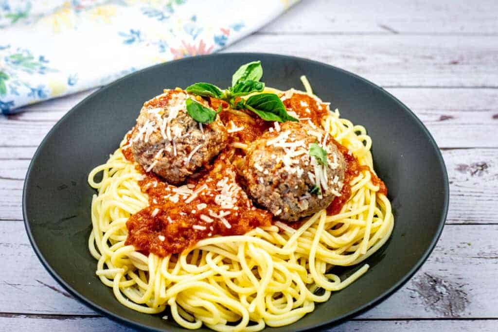 A plate of spaghetti topped with two meatballs, marinara sauce, grated cheese, and basil garnish.