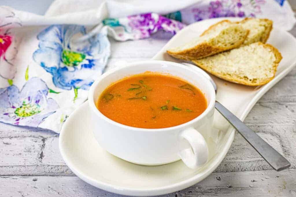 A white cup filled with tomato soup garnished with herbs sits on a matching saucer with a slice of bread.