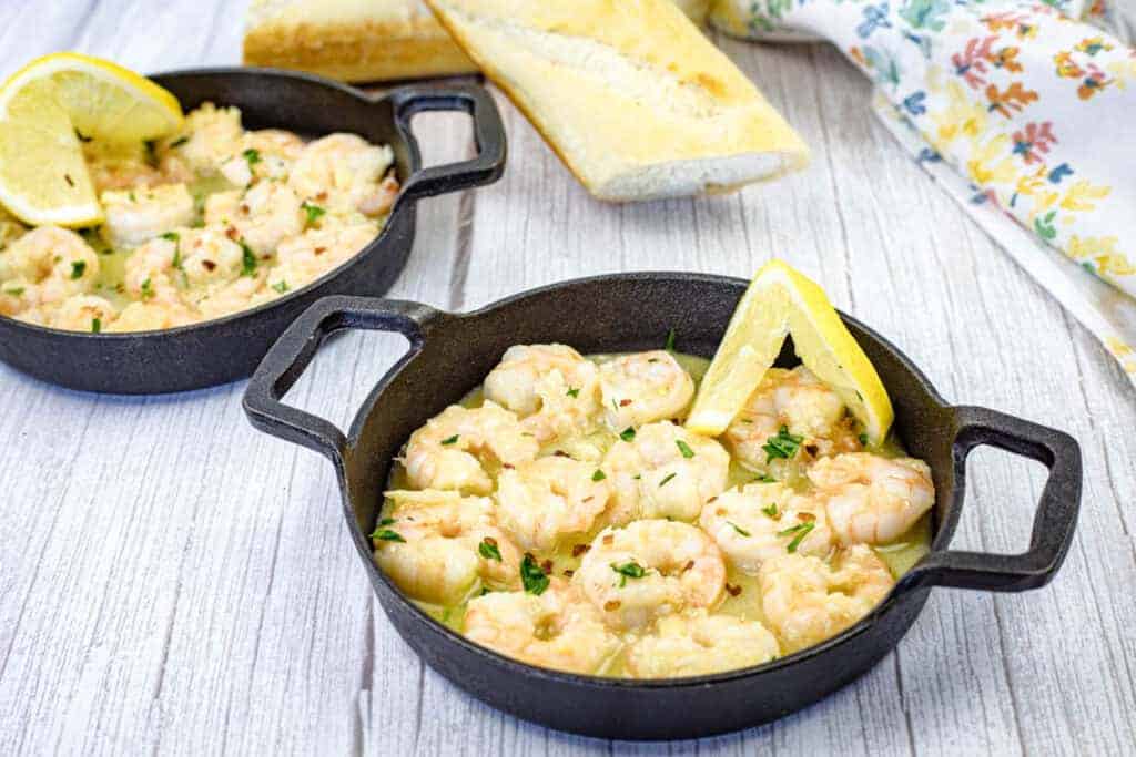 Two cast iron skillets contain shrimp scampi garnished with lemon wedges.