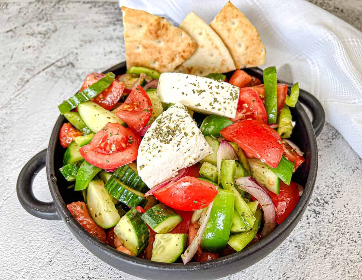 A black bowl contains a Horiatiki salad with blocks of feta cheese, garnished with herbs.