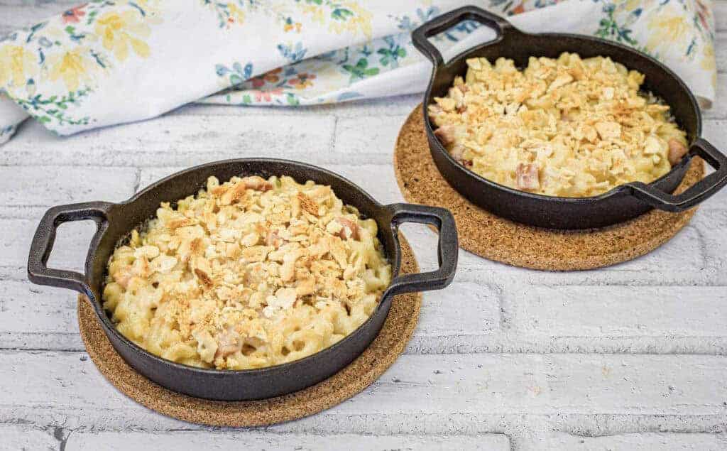 Two black cast iron pans filled with mac and cheese sit on cork trivets on a white wooden surface.