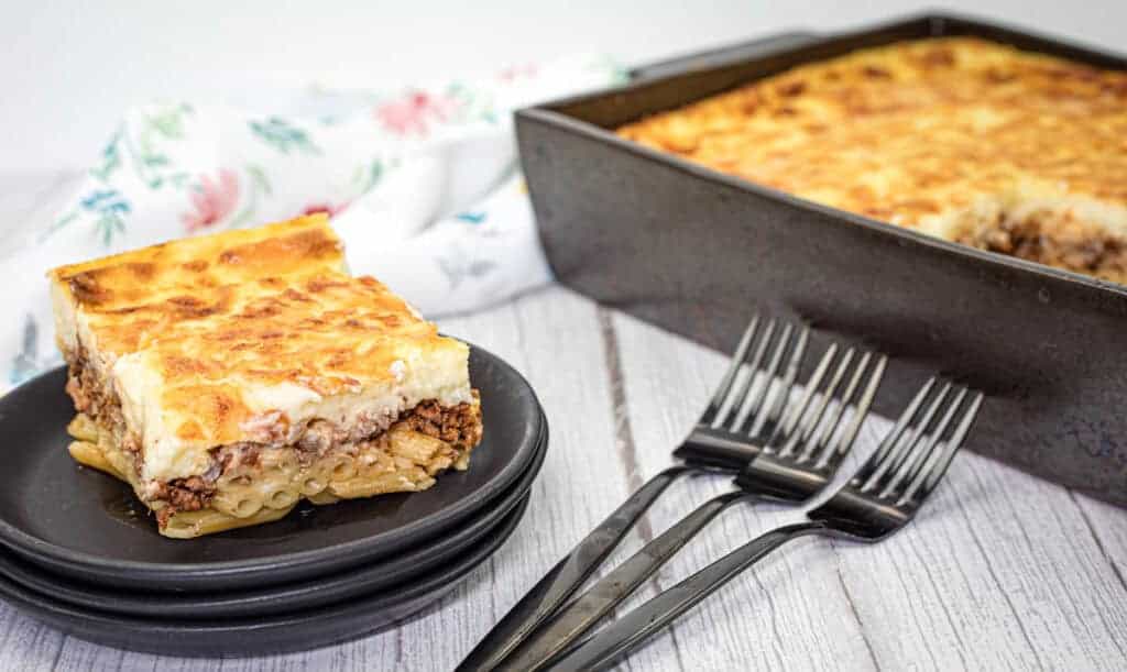 A serving of pastitsio in a black dish is placed in front of a baking tray with more pasta.