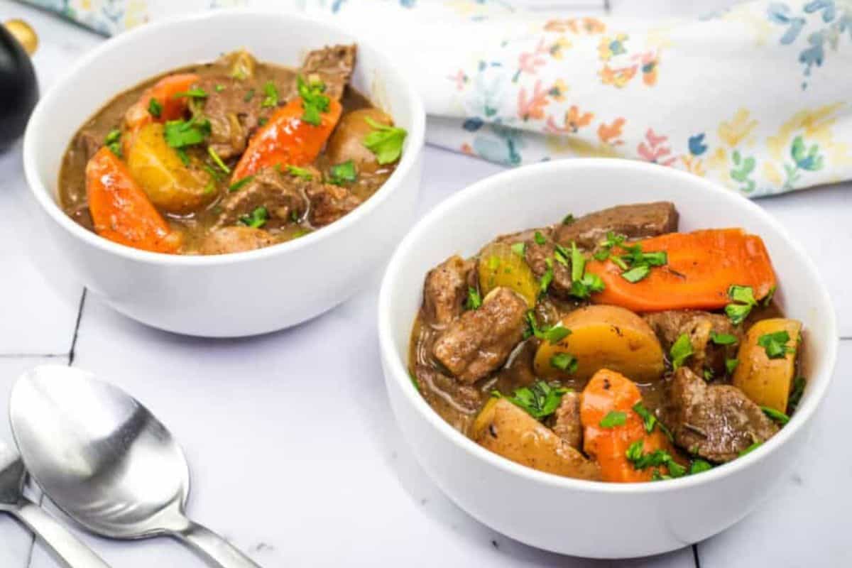 Two bowls of beef stew alongside two silver spoons.