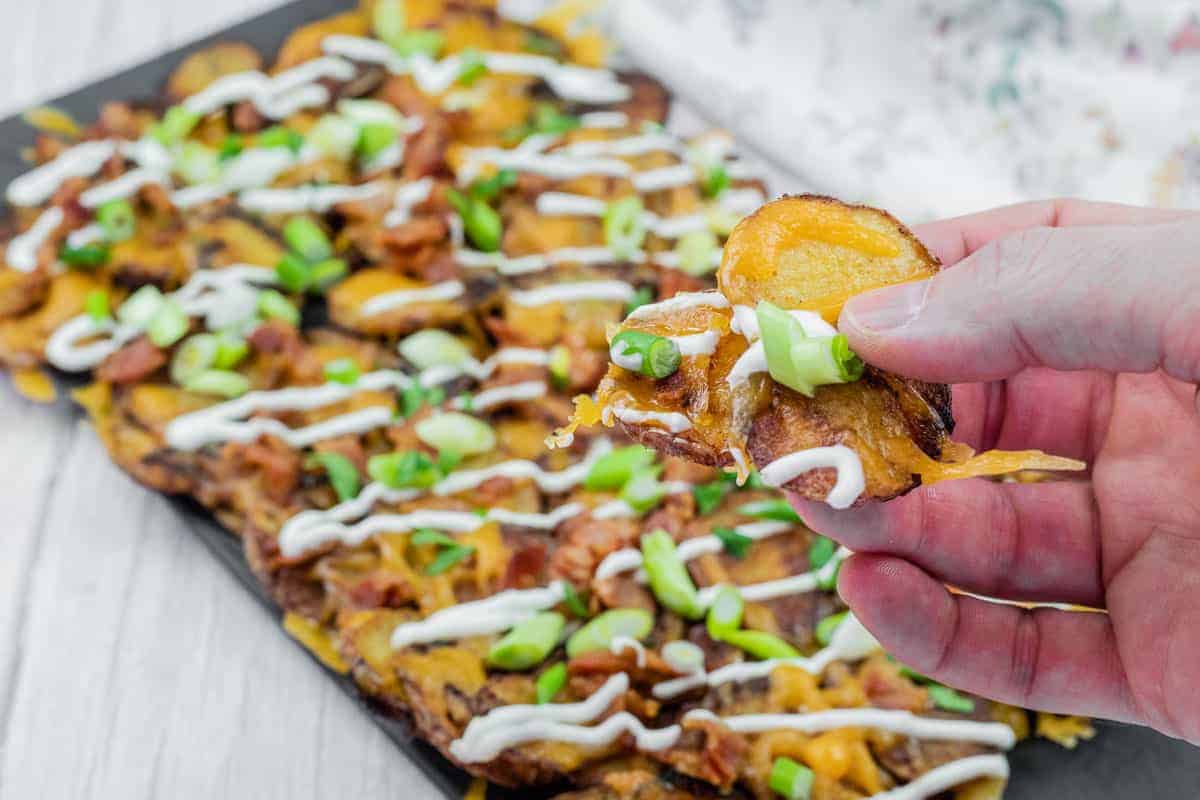 Hand holding a loaded Irish nachos with cheese, bacon, sour cream, and green onions, over a platter of similarly topped slices on a table.