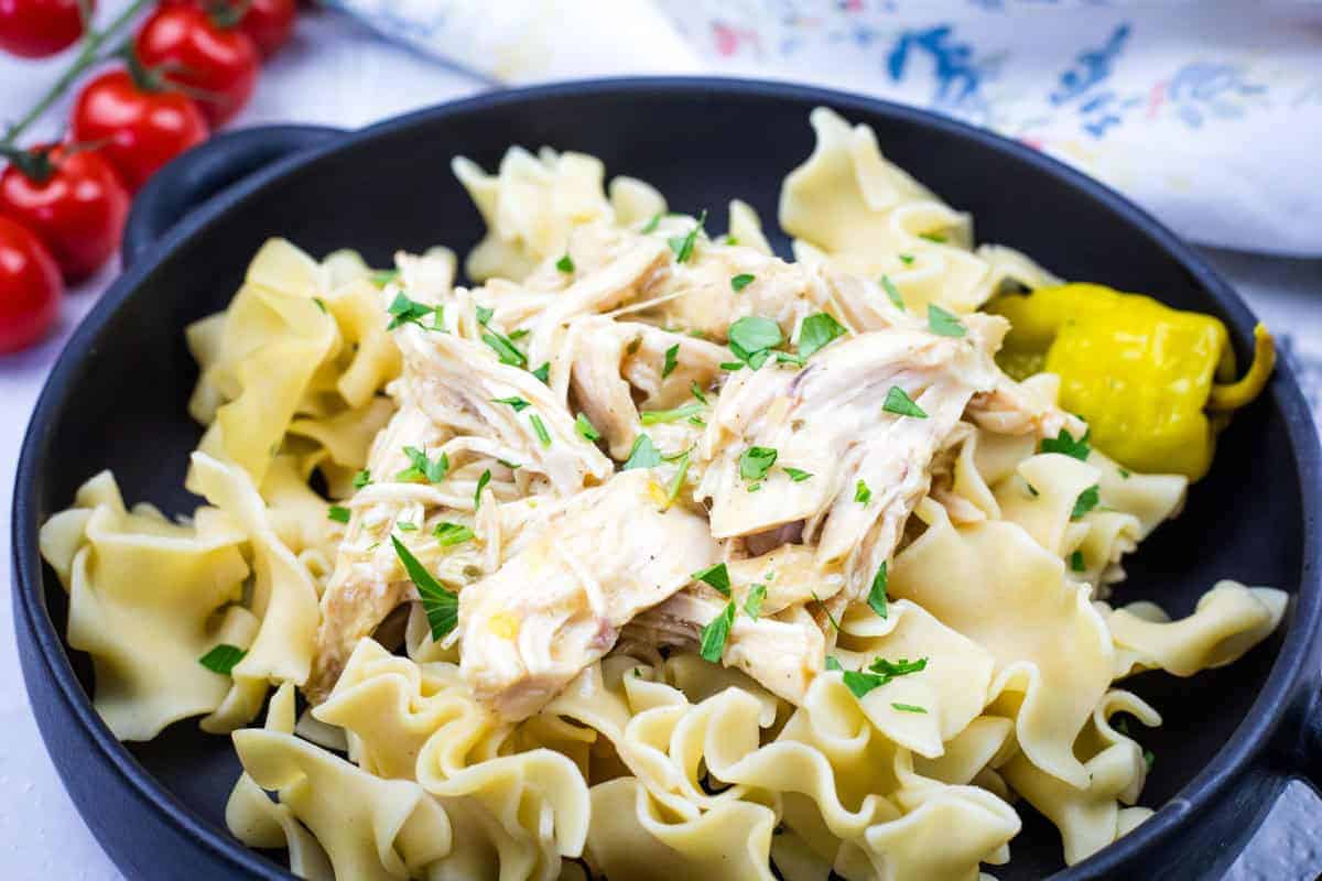 A black bowl containing egg noodles topped with Slow Cooker Mississippi Chicken and garnished with parsley, accompanied by a pickled pepper.
