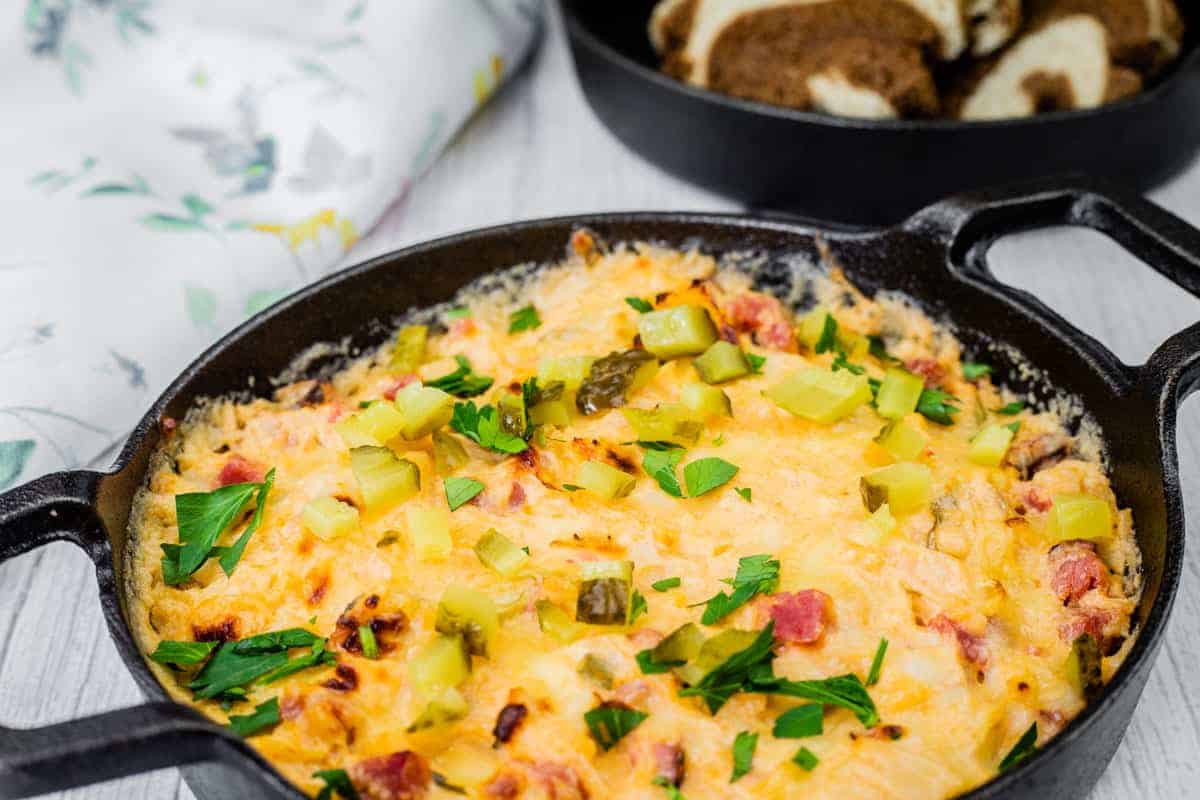 Cheesy, golden-brown Hot Reuben dip in a cast-iron skillet, topped with chopped pickles and fresh parsley, served with slices of marbled rye bread.