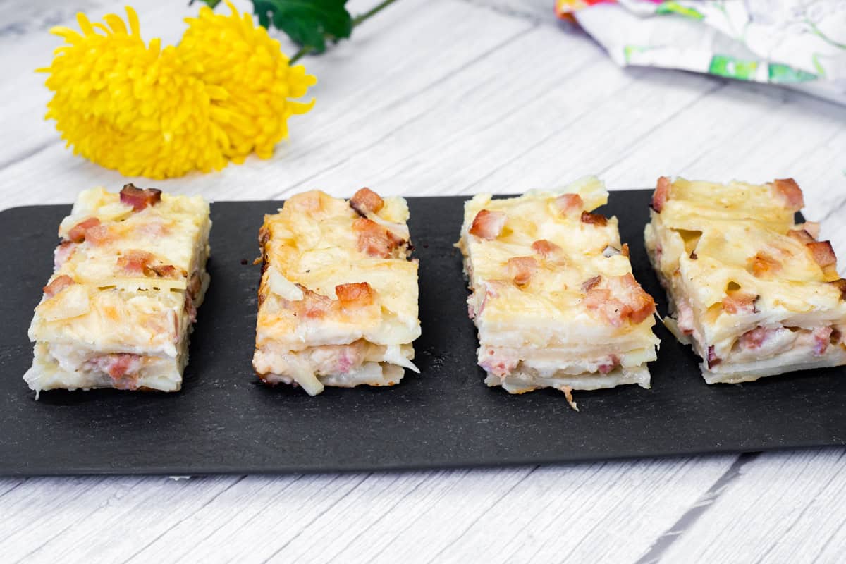 Four slices of Scalloped Potatoes with Ham and Cheese on a black slate board, placed on a light wooden surface.