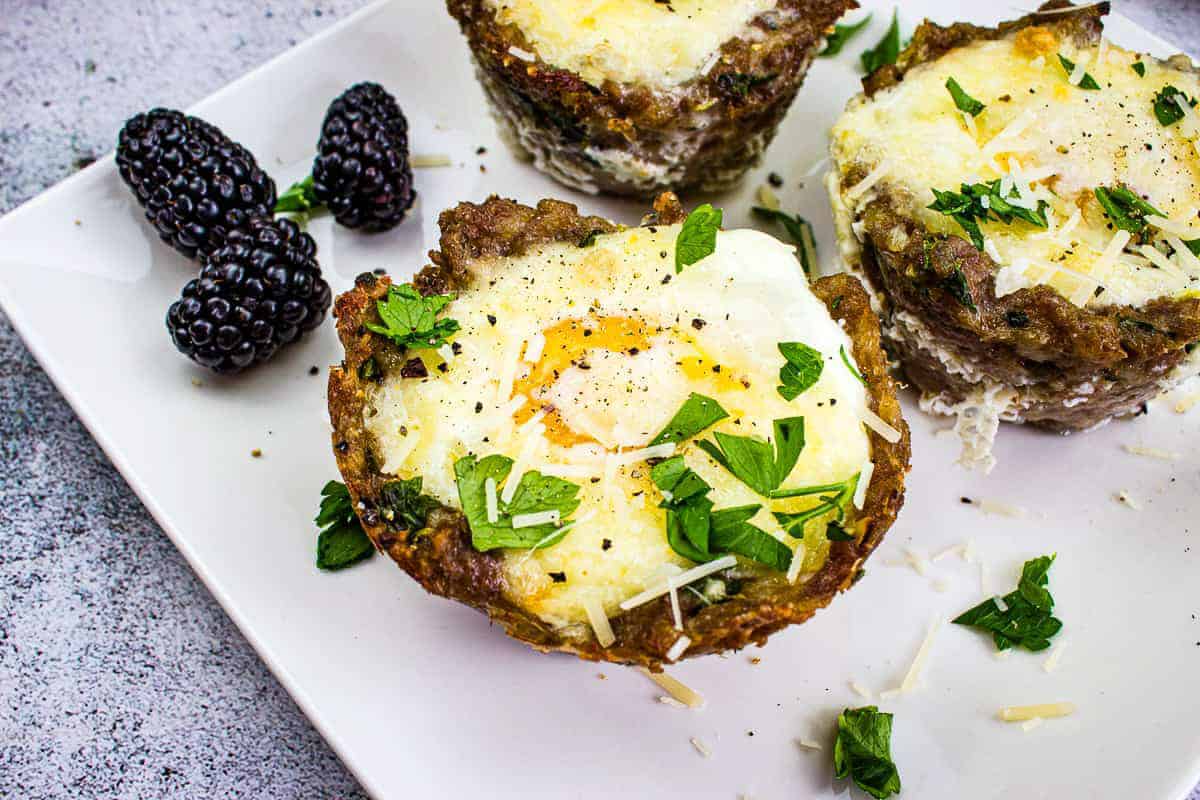 Three baked egg and meat cups topped with herbs and cheese are on a white plate, accompanied by blackberries.
