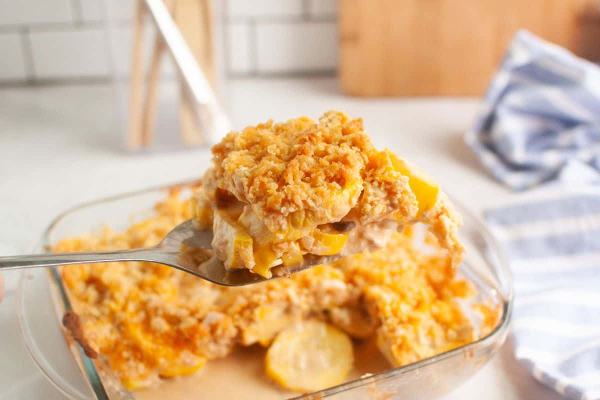 A spoon holds a serving of yellow squash casserole with a crumbly topping above a glass baking dish on a kitchen counter.
