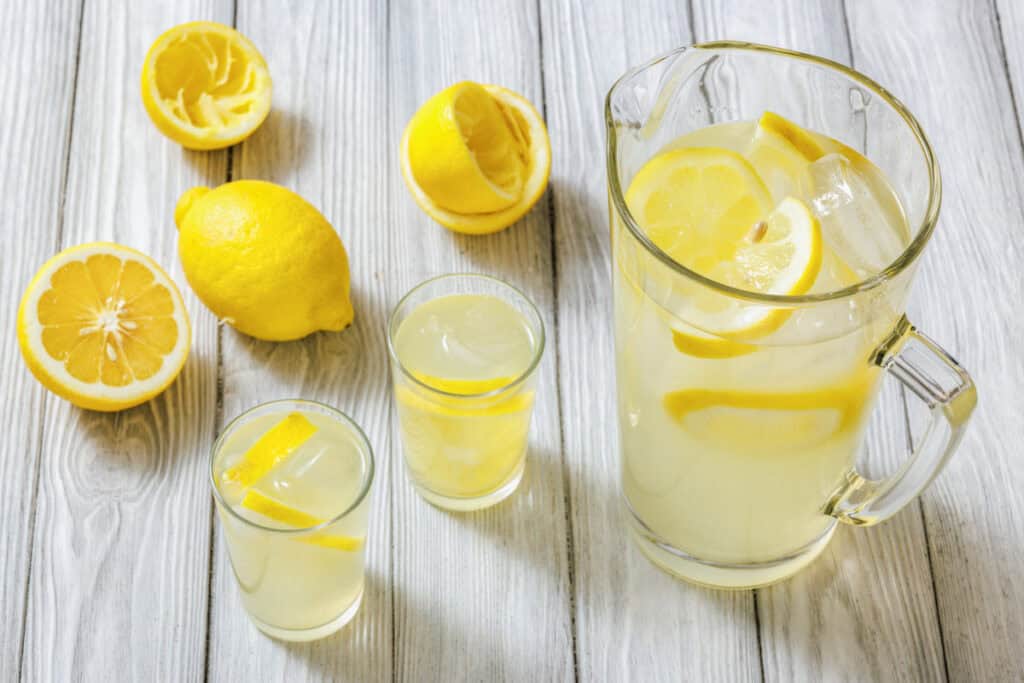 A glass pitcher and two glasses of lemonade with lemon slices, surrounded by whole and halved lemons on a light wooden surface.