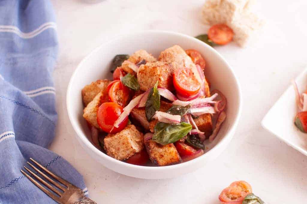 A white bowl filled with panzanella salad, featuring bread cubes, cherry tomatoes, basil leaves, and sliced red onions on a white surface.