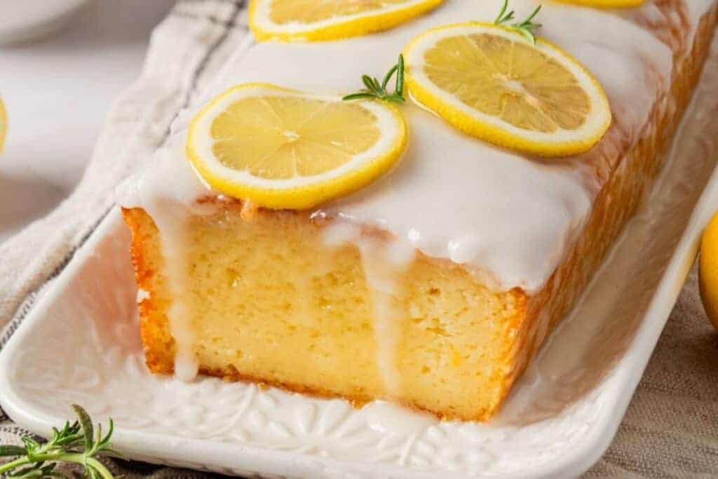 A rectangular lemon loaf cake with white icing drips, topped with lemon slices and herbs, sits on a white platter.