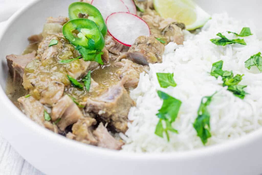 Close-up of a bowl of slow cooker pork chile verde served with white rice, garnished with fresh cilantro, sliced jalapeños, radish slices, and a wedge of lime.