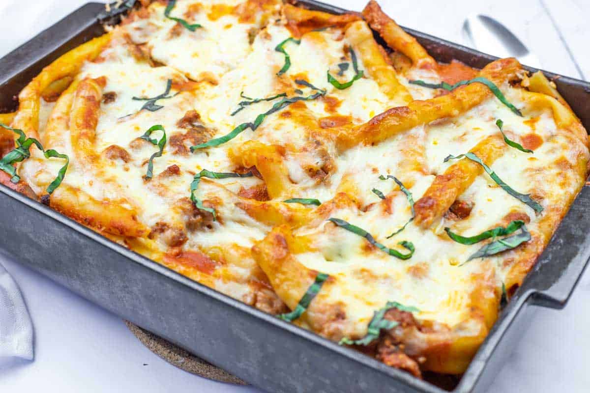Baked Ziti Casserole dish in a rectangular pan topped with melted cheese, tomato sauce, ground meat, and fresh basil strips.