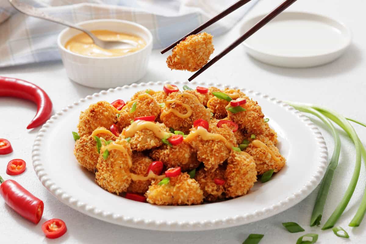 A plate of Bang Bang Chicken Bites garnished with sliced red chili, green onions, and drizzled sauce, with a bowl of dipping sauce in the background.