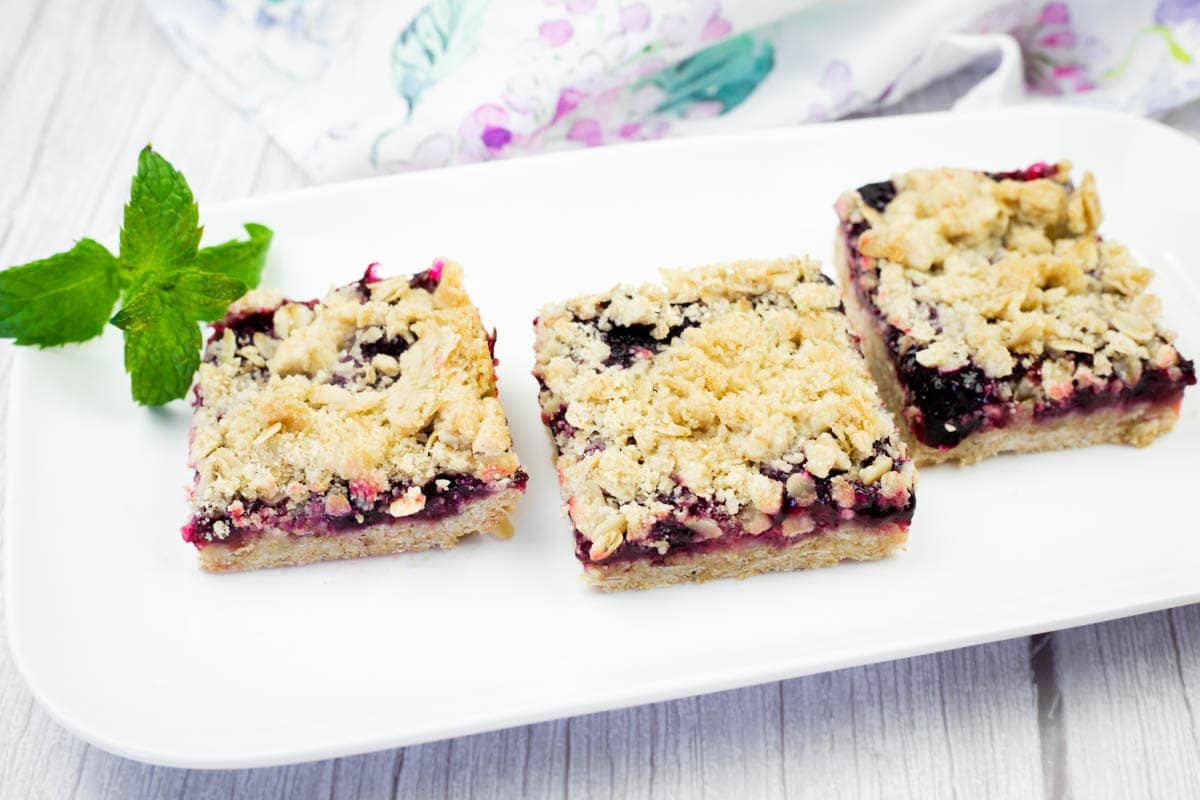 Three square Blueberry-Rhubarb Crumble Bars with golden, crumbly tops and vibrant purple fruit fillings are arranged on a white rectangular platter, garnished with fresh mint.