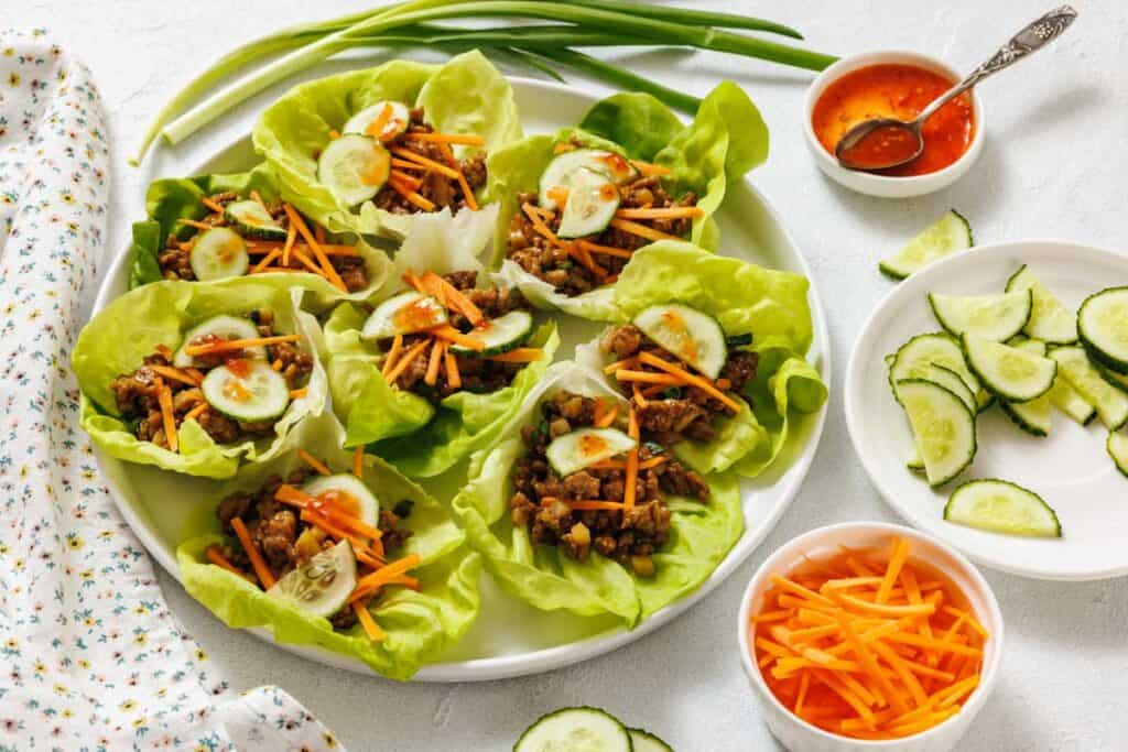 A plate of chicken lettuce wraps filled with shredded carrots and cucumber slices, with bowls of sliced cucumbers, shredded carrots, and dipping sauce on the side.