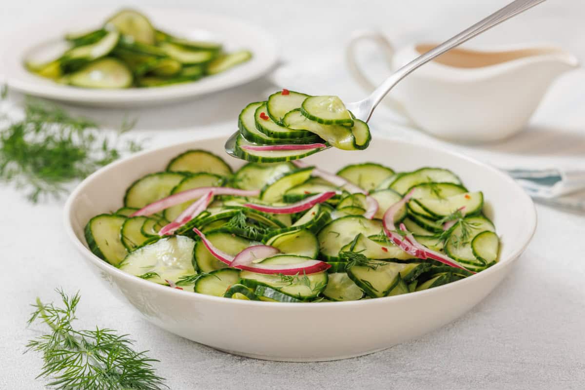A bowl of cucumber salad with red onion and herbs; a spoon is lifting a portion. A plate of salad and a sauce boat are in the background.