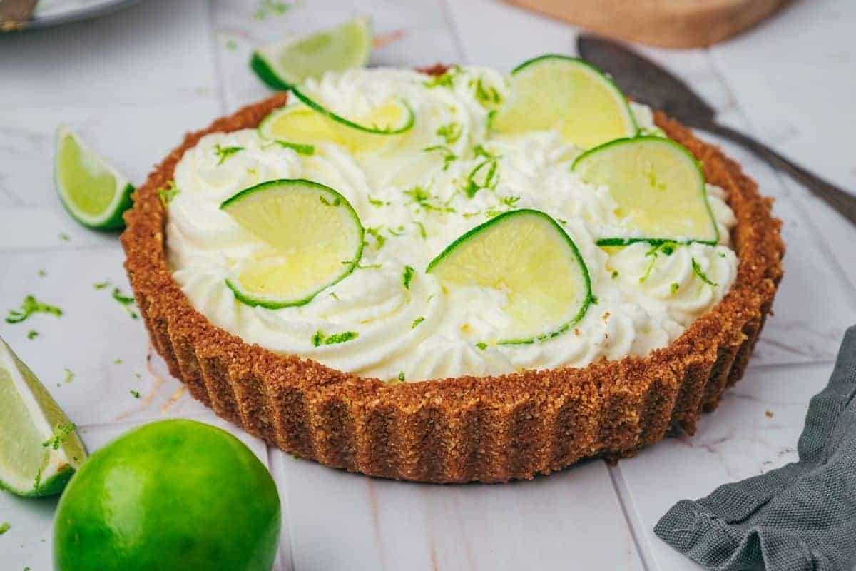 A key lime pie with whipped cream topping, garnished with lime slices and zest, sits on a white surface with additional lime wedges and a knife nearby.