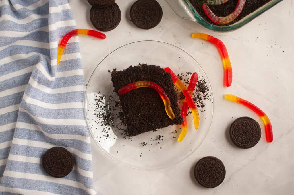 A plate with a slice of dirt cake topped with gummy worms, surrounded by Oreo cookies and a blue-striped cloth on a white surface.