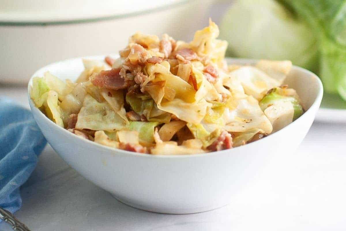 A white bowl filled with fried cabbage with pieces of bacon sits on a white surface with a blue cloth nearby.