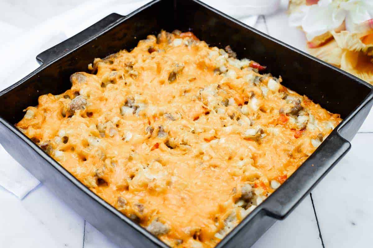 A square black baking dish filled with a baked casserole topped with melted cheese, containing visible pieces of meat and vegetables.