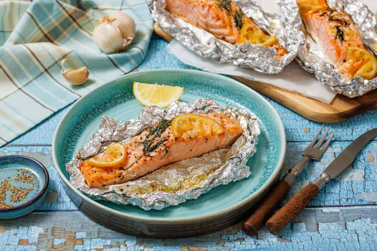 Lemon-Dill Salmon Foil Packets and herbs served in aluminum foil on a blue plate, with utensils and garlic nearby.