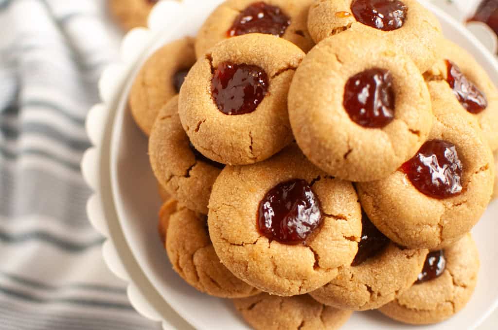 A plate stacked with Peanut butter jelly thumbprint cookies filled with red jam, placed on a white stand next to a striped cloth.