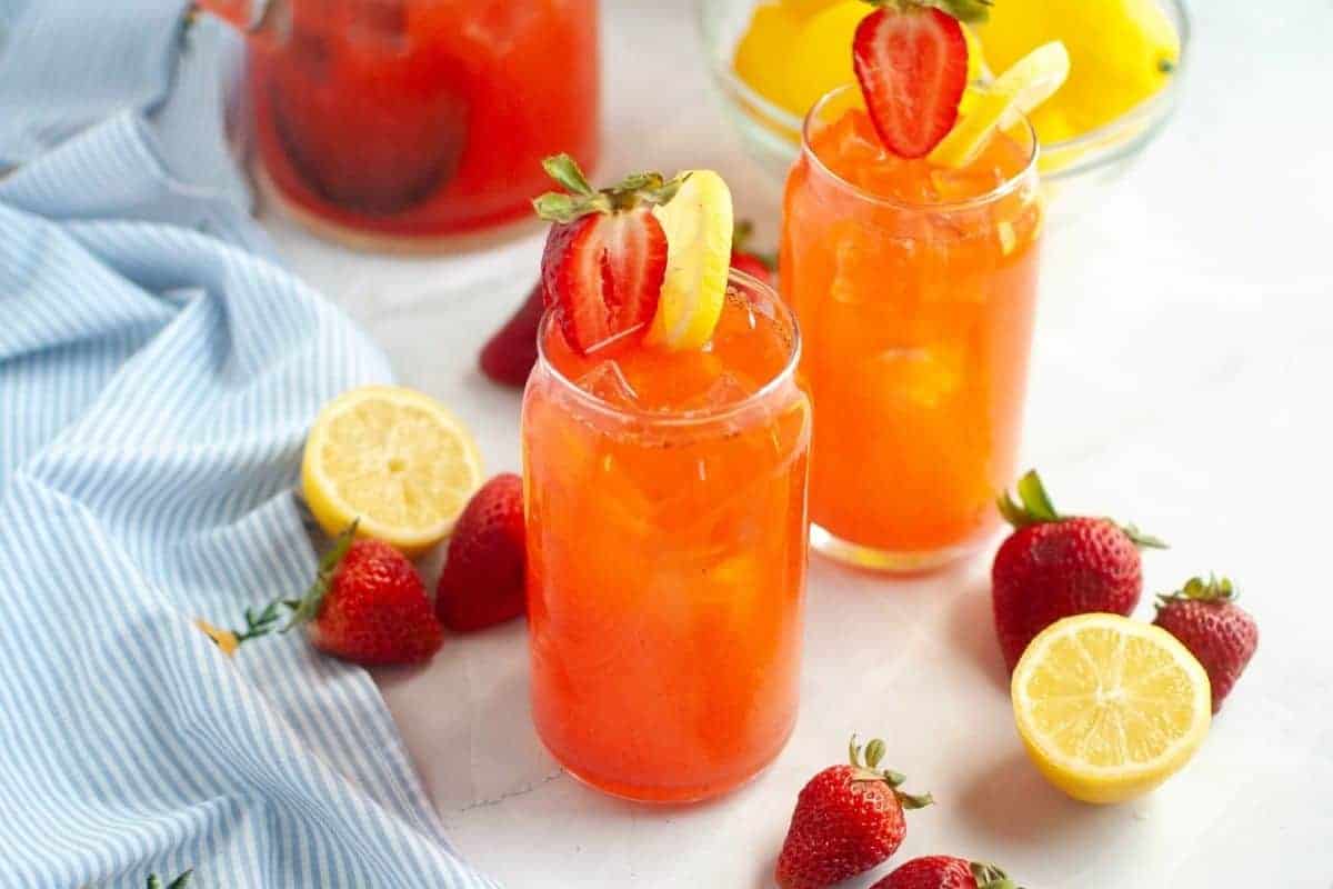 Two glasses of strawberry lemonade with ice, garnished with sliced strawberries and lemons, surrounded by fresh strawberries, lemon halves, and a blue striped cloth.