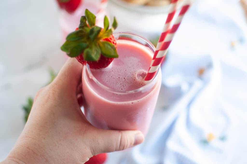 A hand holding a glass of pink strawberry milk with a red and white straw and a strawberry garnish.