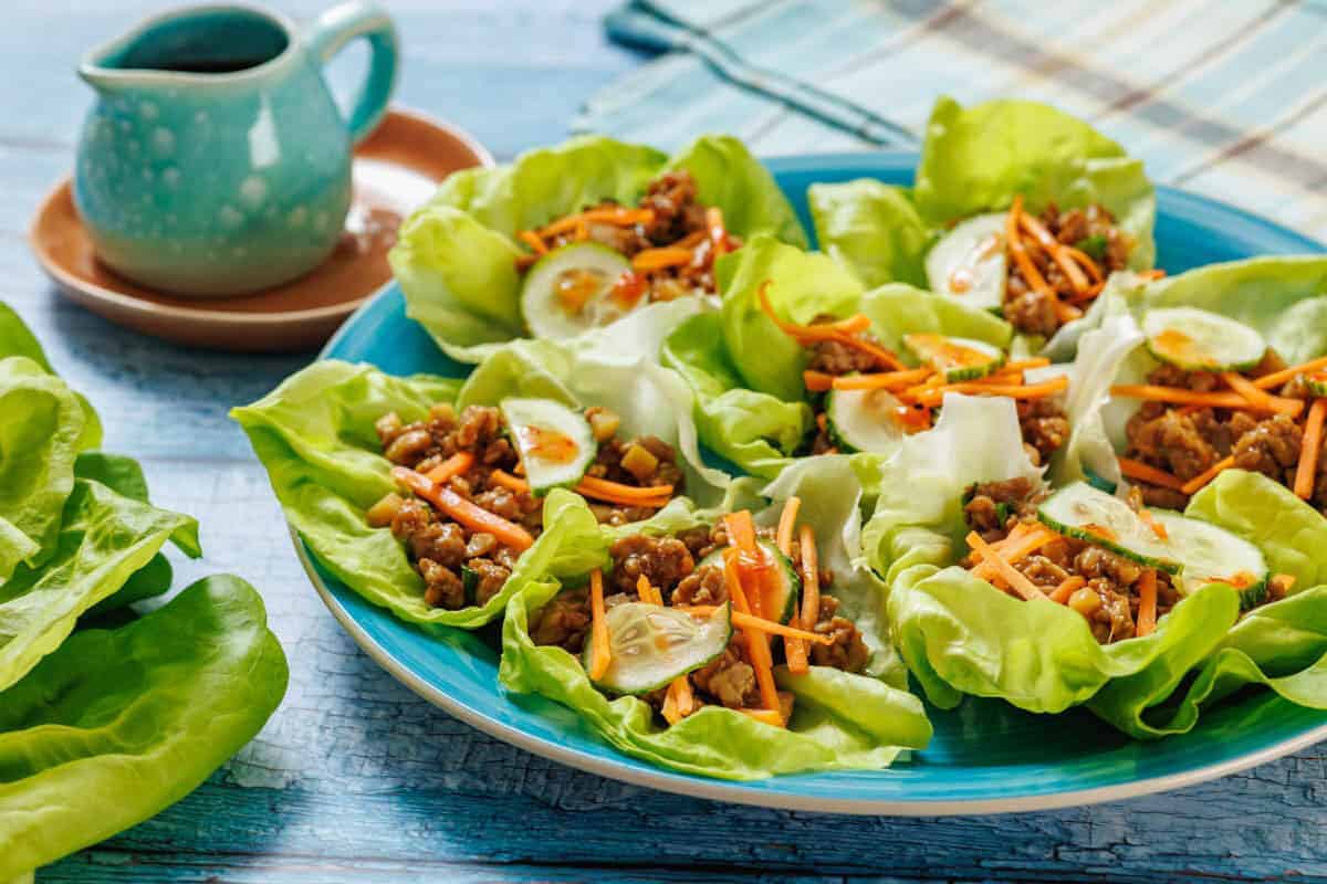 A plate of chicken lettuce wraps filled with minced meat, shredded carrots, and cucumber slices, with a small pitcher of sauce in the background.