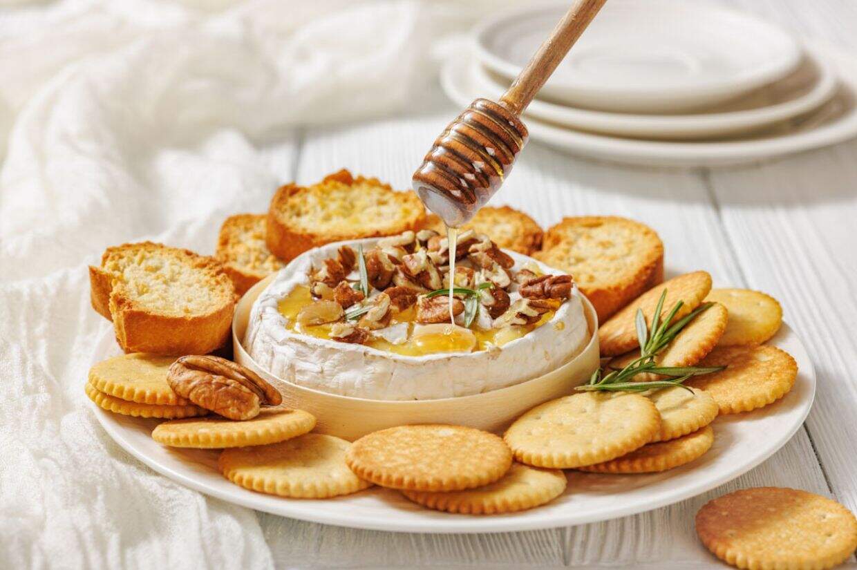A plate with Baked Brie with Honey and Pecans surrounded by crackers and bread slices, with a honey dipper drizzling honey over the cheese.