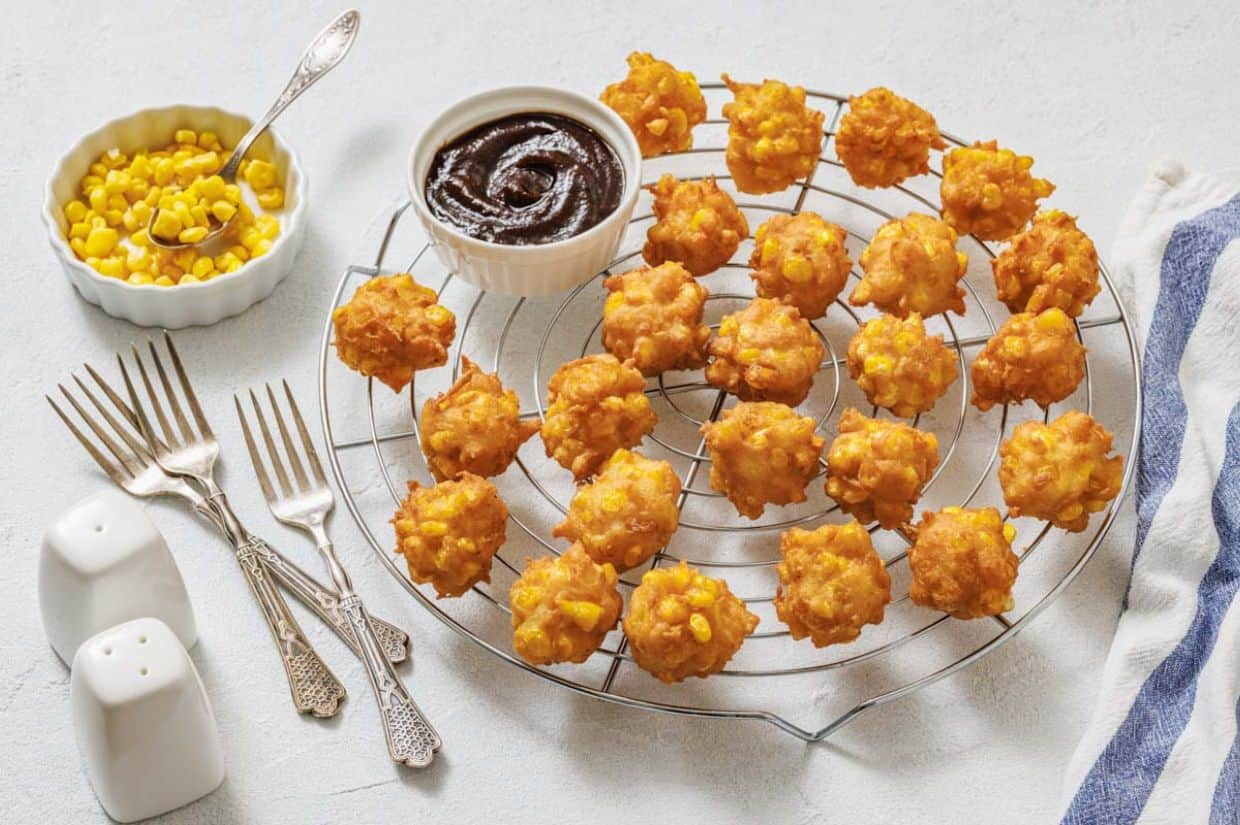A cooling rack with corn nuggets, a bowl of dipping sauce, a bowl of corn, three forks, a spoon, and salt and pepper shakers on a light surface.