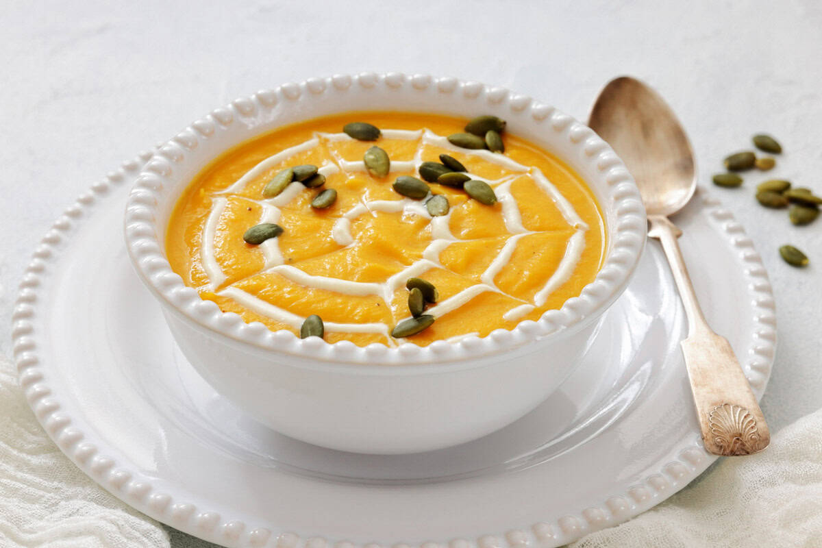 A bowl of creamy pumpkin soup garnished with a swirl of cream in a spiderweb pattern and sprinkled with pumpkin seeds, served with a spoon on a white plate.
