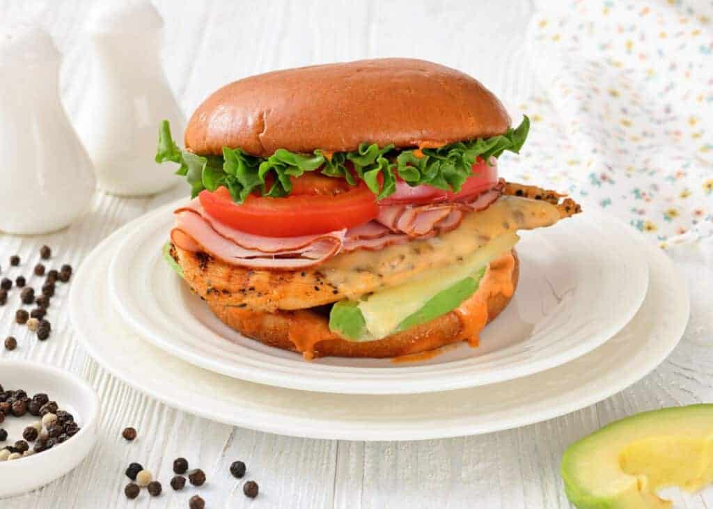 A Grilled California Chicken Burger served on a white plate with peppercorns and a napkin nearby.