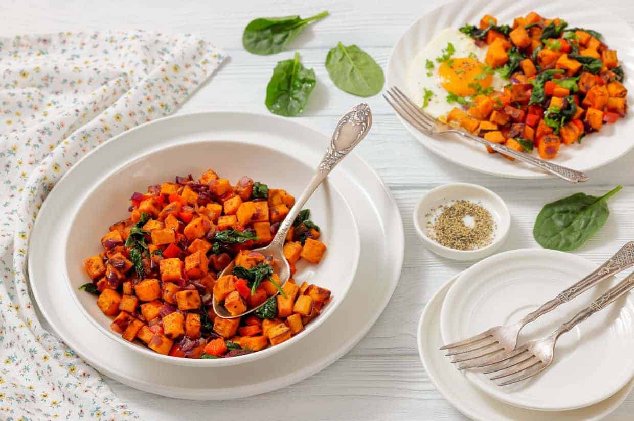 A bowl and plates of sweet potato hash with saut&eacute;ed spinach and onions, served on a white table with forks, a napkin, and a small bowl of pepper.