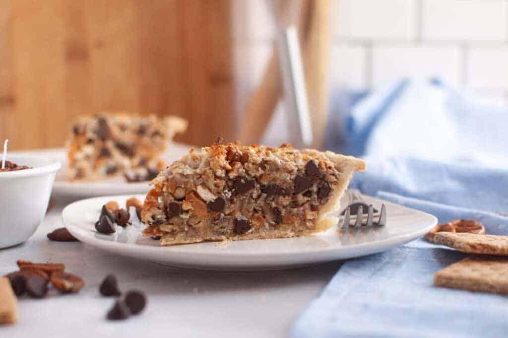 A slice of Texas Trash Pie on a white plate with a fork, with another pie slice and a bowl in the background.
